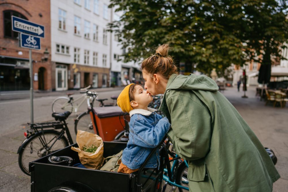 Ung kvinne kysser et barn ca 3 år som sitter foran i en sykkelvogn