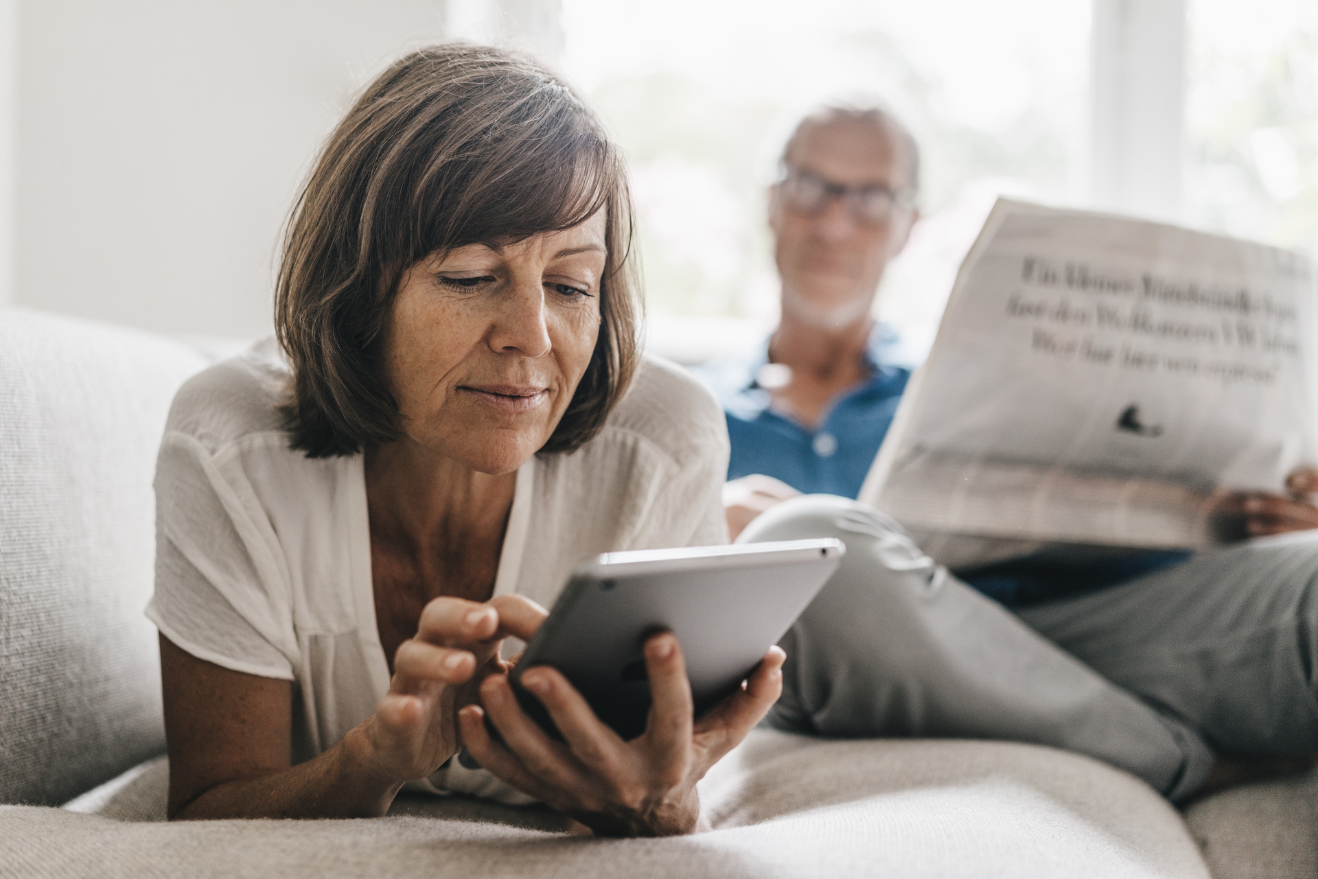 Kvinne ligger på sofaen og ser på en Ipad mannen sitter i bakgrunnen og leser avisen